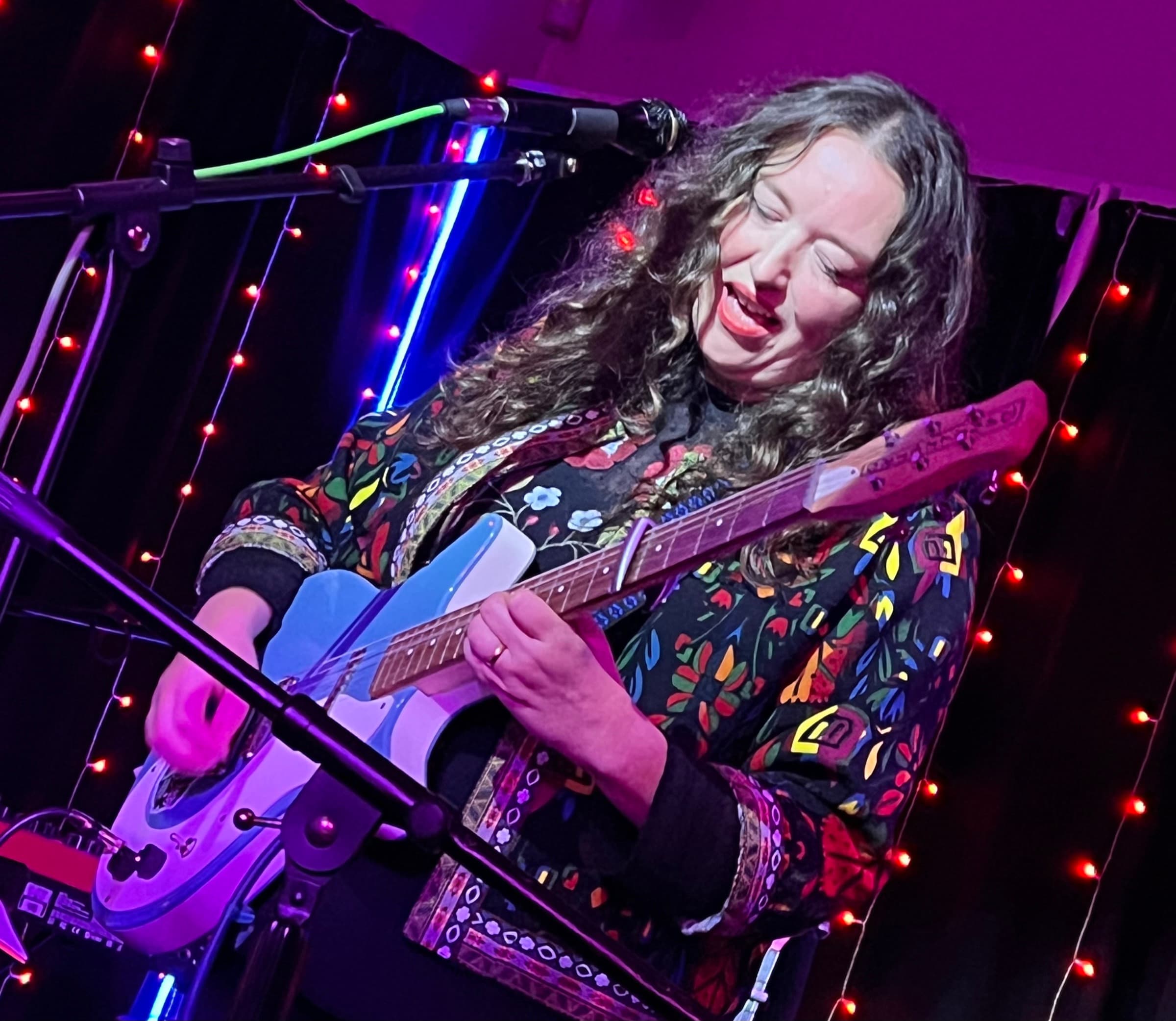 Kathryn Williams: a guitarist with long dark curls playing a pale blue electric guitar under pink light